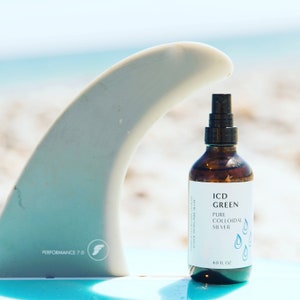 Puede incluir: Una botella de vidrio transparente con un pulverizador negro, que contiene ICD Green Pure Colloidal Silver. La botella está sentada sobre una tabla de surf azul con una aleta blanca.