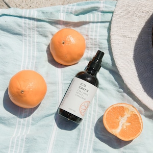 May include: An overhead shot features three oranges, one cut in half, a bottle of ICD GREEN Colloidal Silver + Tangerine, and a white hat on a striped blue towel. The bottle has a black spray top and a label with text and a citrus slice graphic.