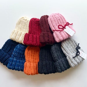 May include: A collection of ten knitted hats in various colors, including beige, burgundy, pink, gray, navy blue, orange, and black. The hats are arranged in a semi-circle, with the pink hat at the top.