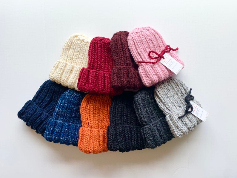 May include: A collection of ten knitted hats in various colors, including beige, burgundy, pink, gray, navy blue, orange, and black. The hats are arranged in a semi-circle, with the pink hat at the top.