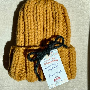 May include: A mustard yellow hand-knitted cuff hat with a black ribbon tie. The hat is made in Arizona and is labeled "7SistersShop Hand Knit Cuff Hat Adult S/M".