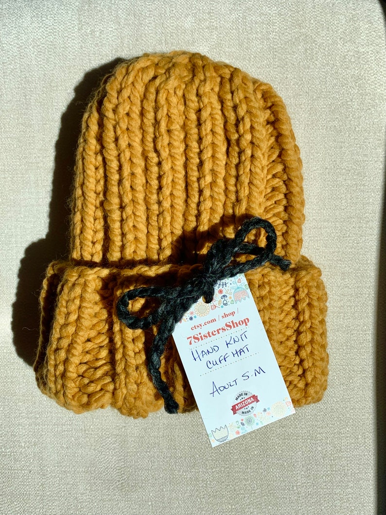 May include: A mustard yellow hand-knitted cuff hat with a black ribbon tie. The hat is made in Arizona and is labeled "7SistersShop Hand Knit Cuff Hat Adult S/M".
