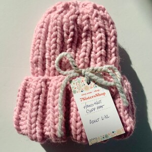 May include: A pink hand-knitted cuff hat with a white ribbon and a tag that reads "etsy.com/shop/7SistersShop Hand Knit Cuff Hat Adult L-XL".
