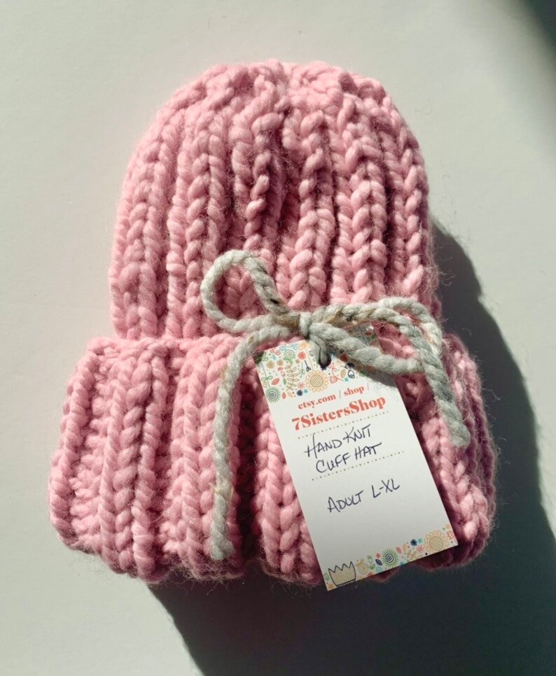 May include: A pink hand-knitted cuff hat with a white ribbon and a tag that reads "etsy.com/shop/7SistersShop Hand Knit Cuff Hat Adult L-XL".