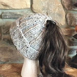 May include: A gray knitted beanie with a ponytail hole. The beanie is made with a textured stitch pattern.