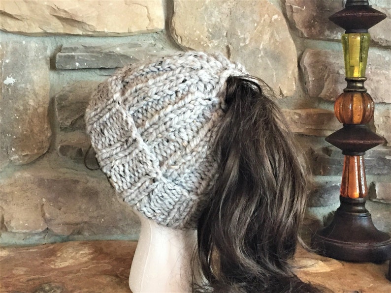 May include: A gray knitted beanie with a ponytail hole. The beanie is made with a textured stitch pattern.