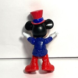 Op de afbeelding: Een Mickey Mouse-beeldje, van achteren gezien, gekleed in een blauwe jas, rode broek en een rode hoge hoed met een blauwe band. De figuur heeft zwarte oren, witte handschoenen en de armen zijn uitgestrekt.