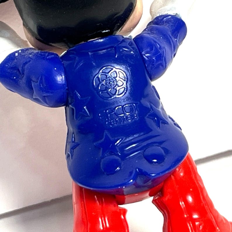 Op de afbeelding: Een close-up van een vintage speelgoedfiguur, met een blauwe jas versierd met sterren en een bloemenembleem. De figuur heeft rode benen en witte handschoenen. De tekst "FCO" is zichtbaar op de jas.