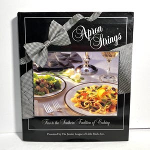 May include: A cookbook titled "Apron Strings" with a black cover and a bow design. The cover features images of food, wine glasses, and text that reads "Ties to the Southern Tradition of Cooking."