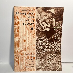 May include: A music instruction book titled Stylings for Bluegrass and Country Guitar, compiled and arranged by Jerry Silverman. The cover features a sepia-toned photograph of a person wearing a cowboy hat and playing an acoustic guitar outdoors.