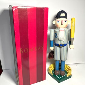Könnte beinhalten: Ein Nussknacker eines Baseballspielers aus Holz mit blau-weiß gestreifter Uniform, der einen gelben Schläger und einen braunen Handschuh hält. Der Nussknacker steht auf einer grün-gelben Stadionbasis. Links ist eine rot-rosa gestreifte Box mit dem Wort "BOMBAY".