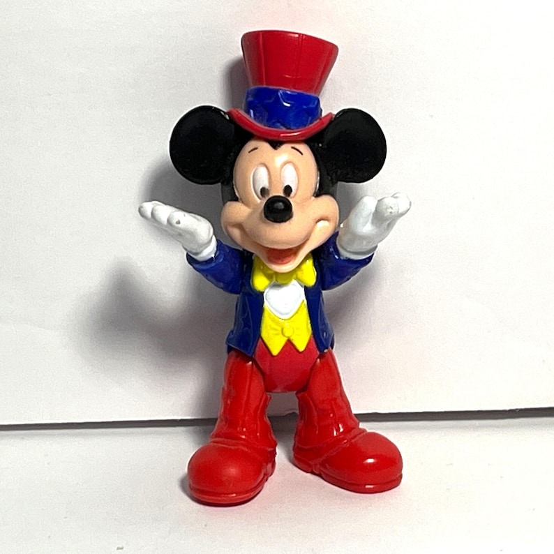 Op de afbeelding: Een Mickey Mouse-beeldje in een goochelaarsoutfit. Het beeldje draagt een rode hoge hoed met een blauwe band, een blauwe jas, een gele vest, een rode broek en rode schoenen. Mickey's witte gehandschoende handen zijn uitgestrekt.