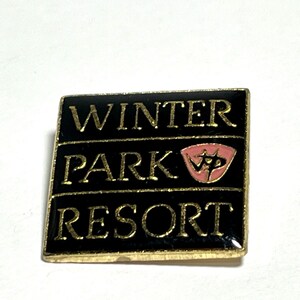 May include: A black and gold enamel pin with the text "WINTER PARK RESORT" in gold lettering. A pink and gold shield with the letters "WP" is in the center of the pin.