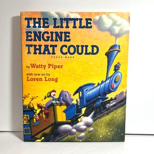 Könnte beinhalten: Ein Buchcover mit gelbem Hintergrund und einer blauen Lokomotive, die einen Waggon mit einem Clown und einem Elefanten zieht. Der Titel des Buches lautet "The Little Engine That Could" von Watty Piper mit neuen Illustrationen von Loren Long.