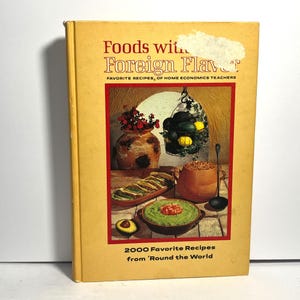 May include: A vintage cookbook titled "Foods with Foreign Flavor" with a yellow cover. The cover features a colorful illustration of various dishes and ingredients, including a bowl of guacamole, a wooden tray of food, and a decorative vase. The book's title and subtitle are displayed prominently.