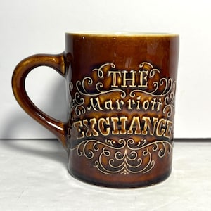 Może przedstawiać: Brązowy ceramiczny kubek z zakrzywionym uchwytem. Kubek zawiera słowa "THE Marriott EXCHANGE" w ozdobnej, kremowej czcionce. Kubek ma błyszczące wykończenie i kremowe wnętrze.