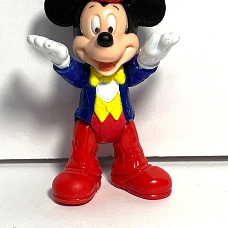 Op de afbeelding: Een Mickey Mouse-beeldje in een klassieke outfit. Het figuur heeft een rode hoed, een rode broek en rode schoenen. De jas is blauw met een gele gilet en een vlinderdas. Mickey's witte handschoenen zijn zichtbaar terwijl hij zijn armen uitstrekt.