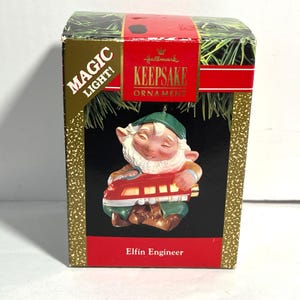 May include: A Hallmark Keepsake ornament box featuring an elf engineer. The box has a red band and green foliage. The ornament shows a smiling elf in a green hat and brown jacket, holding a red train. Text on the box reads "Magic Light!" and "Elfin Engineer."