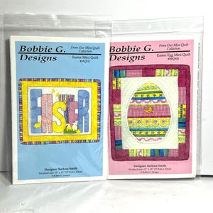 Könnte beinhalten: Zwei Oster-Mini-Quilt-Muster von Bobbie G. Designs. Das erste Muster zeigt das Wort "Ostern" in bunten Buchstaben auf gelbem Hintergrund. Das zweite Muster zeigt ein rosa-weißes Ei mit einem gelben Vogel darin. Beide Muster sind 11 Zoll mal 13 Zoll groß.