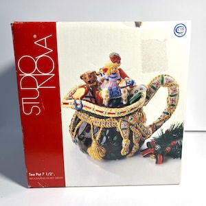 May include: Studio Nova tea pot in a box. The tea pot is shaped like a bag with a handle and is decorated with a Santa Claus figure, a teddy bear, and other toys. The box reads "Tea Pot 7 1/2", Woodland Holly Santa."