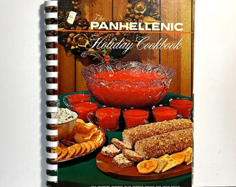 May include: The Panhellenic Holiday Cookbook with a spiral binding. The cover features a festive holiday spread with a large punch bowl, glasses, and various food items. The text reads "900 Favorite Recipes Plus Festive Menus for Year Round Holiday Entertaining."