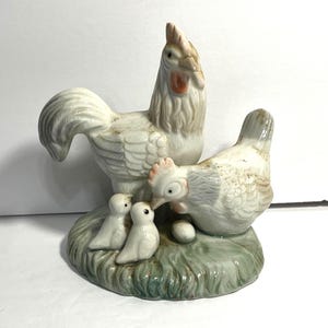 May include: A ceramic figurine depicting a rooster, hen, and two chicks on a grassy base. The rooster stands tall, while the hen appears to be looking at an egg. The figurines are primarily white with touches of red and brown.