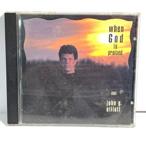 May include: A compact disc case with a sunset background and a man in a black shirt. The text on the front reads "when God is praised" and "john g. elliott".