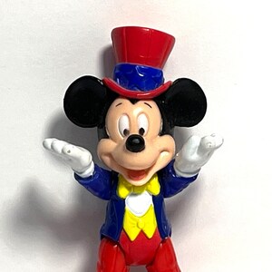 Op de afbeelding: Een Mickey Mouse-beeldje in een goochelaarsoutfit. Het figuur heeft een rode hoge hoed, een blauwe jas, een rode broek en een gele vlinderdas. Mickey's zwarte oren en witte handschoenen zijn zichtbaar.