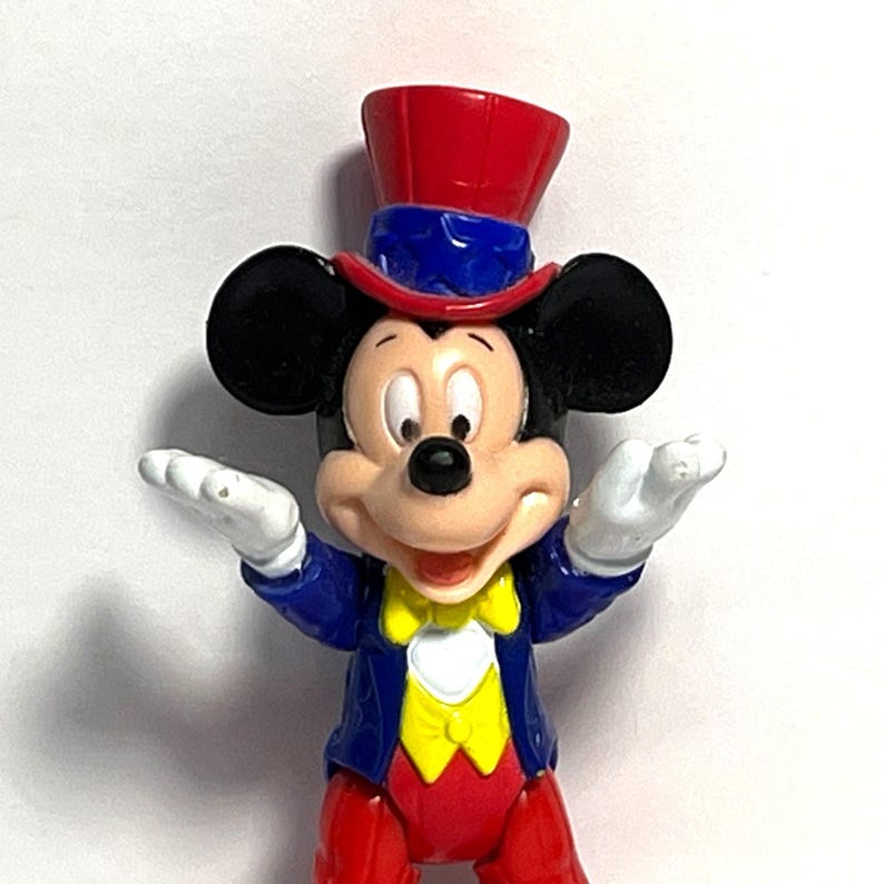Op de afbeelding: Een Mickey Mouse-beeldje in een goochelaarsoutfit. Het figuur heeft een rode hoge hoed, een blauwe jas, een rode broek en een gele vlinderdas. Mickey's zwarte oren en witte handschoenen zijn zichtbaar.