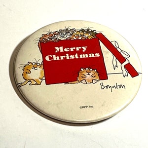 Op de afbeelding: Een ronde knop met een cartoonillustratie van een rode cadeaudoos die overloopt met katten. De doos heeft een wit lint en de tekst "Merry Christmas". De handtekening van de kunstenaar "Benton" staat in de rechterbenedenhoek.