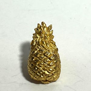 Puede incluir: Un broche de piña de color dorado con un diseño detallado. La piña es una fruta tropical popular y un símbolo de hospitalidad.
