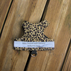 May include: A baby cheetah print dog harness with a reflective strip. The harness features a brown and black spotted pattern, a black strap, and a metal ring. The text "Baby Cheetah with Optional Reflective Strip" is visible.