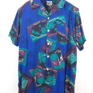 May include: A blue short-sleeved button-down shirt with a colorful print of maps, airplanes, and travel-related text. The shirt has a collar and a pocket on the left side. The text on the shirt includes "First Class", "Baggage", "Steamshipline", "State Room", "Dollars", and "Ken Warren's Finest".