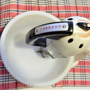 May include: A vintage Kenmore stand mixer in white with a black handle and speed control dial. The mixer is positioned over a white bowl, set against a red and white plaid cloth.