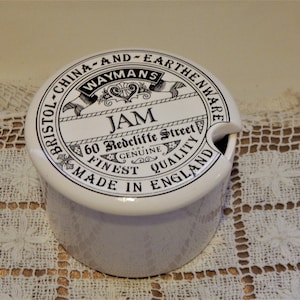 Puede incluir: Tarro de mermelada de cerámica blanca con tapa. La tapa tiene texto negro que dice "WAYMANS JAM" y "Made in England". El tarro tiene una pequeña abertura para una cuchara. Sobre un mantel de encaje blanco.