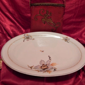 Könnte beinhalten: Eine ovale Servierplatte mit floralen Designs in Grau-, Beige- und Brauntönen. Die Platte hat einen goldenen Rand und ist vor einem roten Hintergrund platziert. Ein roter dekorativer Behälter mit einem Rentier-Design befindet sich im Hintergrund.