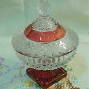 May include: A clear glass candy dish with a lid, featuring a diamond pattern and a red band around the middle. The dish sits on a square, red base with a gold trim. The lid has a decorative knob.