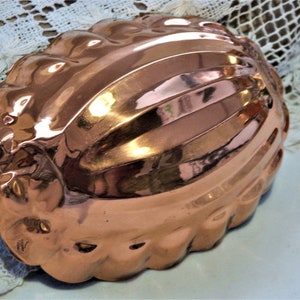 Könnte beinhalten: Eine kupferfarbene, ovale Form mit gewelltem Rand und gerilltem Design. Der Vintage-Küchenartikel hat eine glänzende, reflektierende Oberfläche und eine kleine Metallschlaufe zum Aufhängen. Ein klassisches Backwerkzeug.