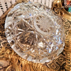 May include: A clear, cut-glass bowl with a starburst design in the center. The bowl has a geometric pattern and is resting on a patterned fabric with gold fringe. The bowl is likely used for decorative purposes.