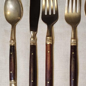 Könnte beinhalten: Ein Set aus Vintage-Besteck, darunter ein Löffel, ein Messer und zwei Gabeln. Die Utensilien haben dunkelbraune Holzgriffe mit Messingakzenten und einem einzelnen weißen Punkt. Das Messer hat eine schwarze Klinge.