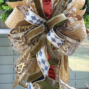 May include: A decorative bow featuring multiple ribbons in gold, olive green, and burnt orange with glittery textures. The design includes a white and blue diamond pattern, metallic gold mesh, and solid velvet ribbons arranged in a layered structure.