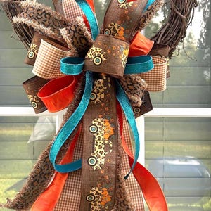 Puede incluir: Un lazo decorativo para corona con una mezcla de cintas de estampado de leopardo marrón, naranja, purpurina turquesa y cuadros vichy marrones. La cinta central marrón muestra un patrón repetitivo de camiones con calabazas naranjas.