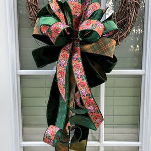 May include: A decorative wreath bow featuring a mix of solid dark green velvet ribbon, a plaid ribbon in shades of green and orange, and a floral patterned ribbon with orange and pink blossoms on an orange background, all tied in a large cluster.