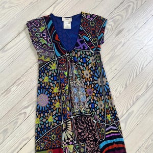 May include: A colorful, patterned dress with a V-neckline. The dress features a patchwork design with various geometric and floral patterns in shades of blue, purple, green, and red. The dress has short sleeves and a blue lining at the neckline.