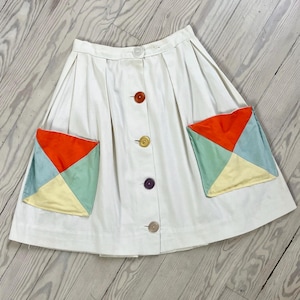 Könnte beinhalten: Ein weißer Rock mit Knopfleiste und zwei großen aufgesetzten Taschen. Die Taschen sind aus einem Patchwork aus gelbem, grünem und orangefarbenem Stoff gefertigt.