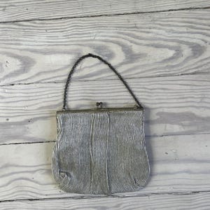 May include: A silver-toned metallic handbag featuring a textured, pleated surface with vertical lines. The bag has a rectangular frame with a metal kiss-lock clasp closure at the top and a thin, dark-colored metal chain strap for carrying.