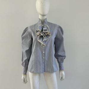 May include: A long-sleeved button-down shirt in a light blue and white pinstripe pattern. The shirt features a high collar, puffed sleeves with buttoned cuffs, and a prominent ruffled lace jabot detail down the center front with white buttons.