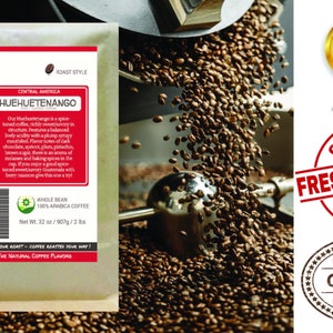 May include: A bag of whole bean coffee labeled "Smokin Beans" with the text "Huehuetenango" and "Central America" printed on the front. The bag is sitting on a table with a coffee roaster in the background.