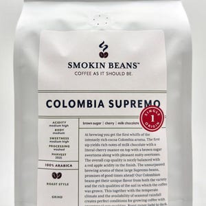 May include: A white coffee bag labeled Smokin Beans Colombia Supremo. The label lists flavor notes of brown sugar, cherry, and milk chocolate, specifies 100% Arabica, and states a net weight of 32 ounces, 2 pounds, or 227 grams on the front.