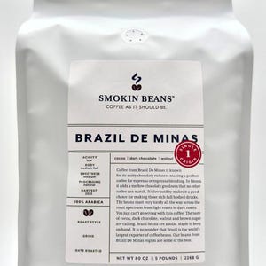May include: A white stand-up pouch of Smokin Beans Brazil De Minas coffee. The label features the brand logo, flavor notes of cocoa, dark chocolate, and walnut, and states a net weight of 80 ounces, 5 pounds, or 2268 grams. It is 100% Arabica coffee.